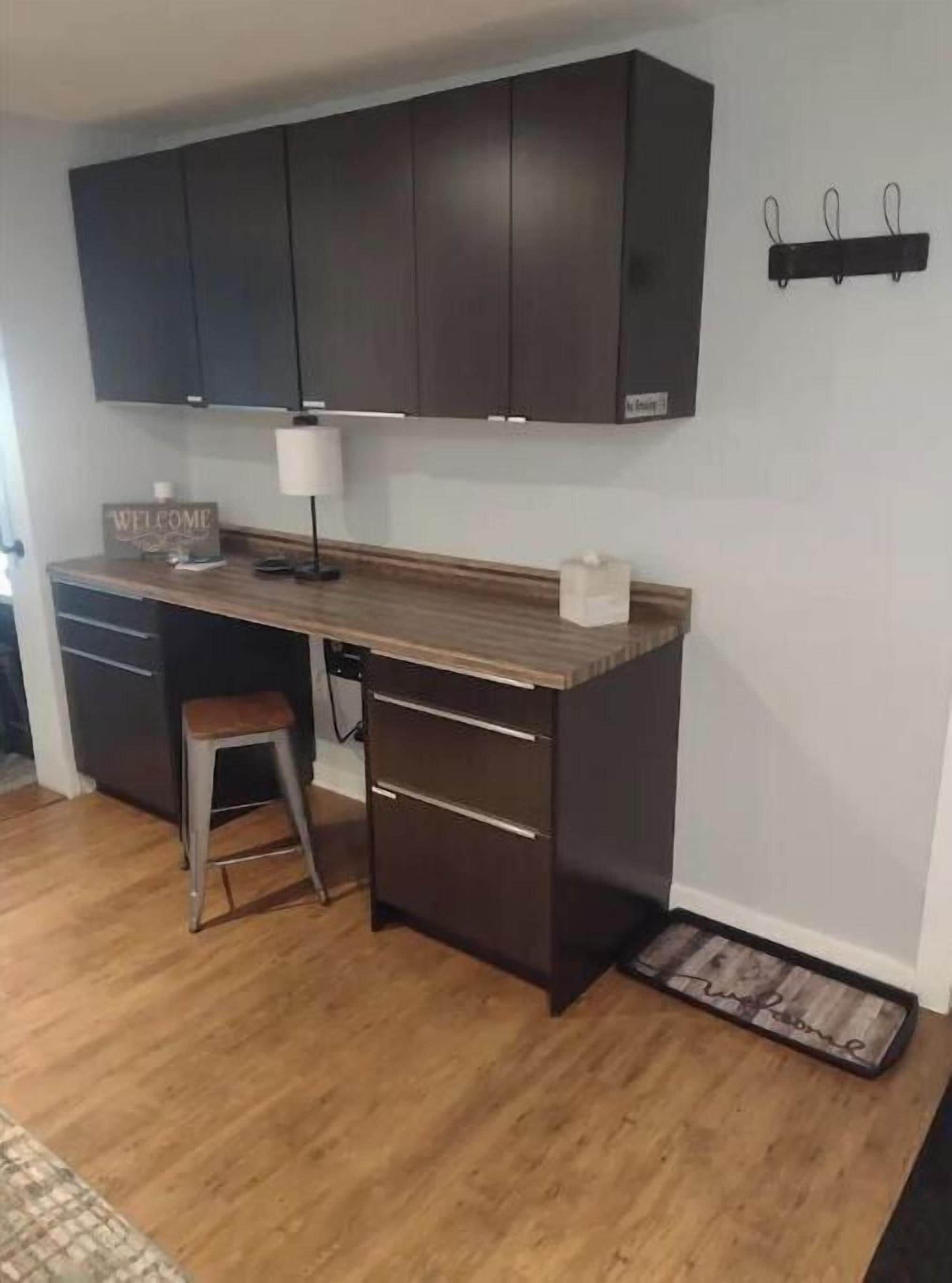 A compact kitchen with dark cabinets, a wooden countertop, a stool, and a welcome sign.