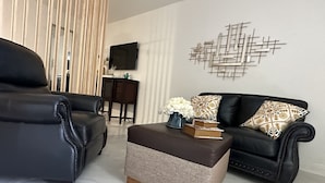 A modern living room with a black leather sofa, a matching ottoman, and a coffee table with decorative items. A TV is mounted on the wall, and there is a decorative wall piece above the sofa. - Kissimmee Studio 16 min from Disney World (Kissimmee)