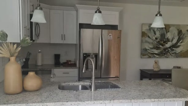 A modern kitchen with white cabinetry, a stainless steel refrigerator, a granite countertop, and two bar stools. - Kissimmee Studio 16 min from Disney World (Kissimmee)