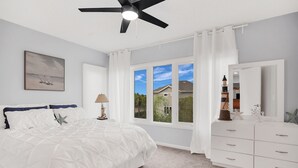 A bedroom with a bed, a ceiling fan, a window with curtains, a dresser, and a painting on the wall.
