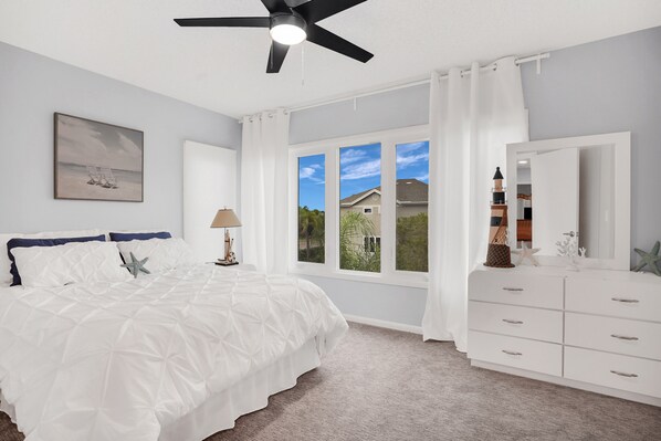 A bedroom with a bed, a ceiling fan, a window with curtains, a dresser, and a painting on the wall.