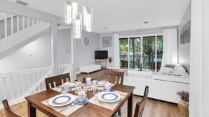 A dining area with a wooden table set for four, a white staircase, and a view of greenery through large windows.