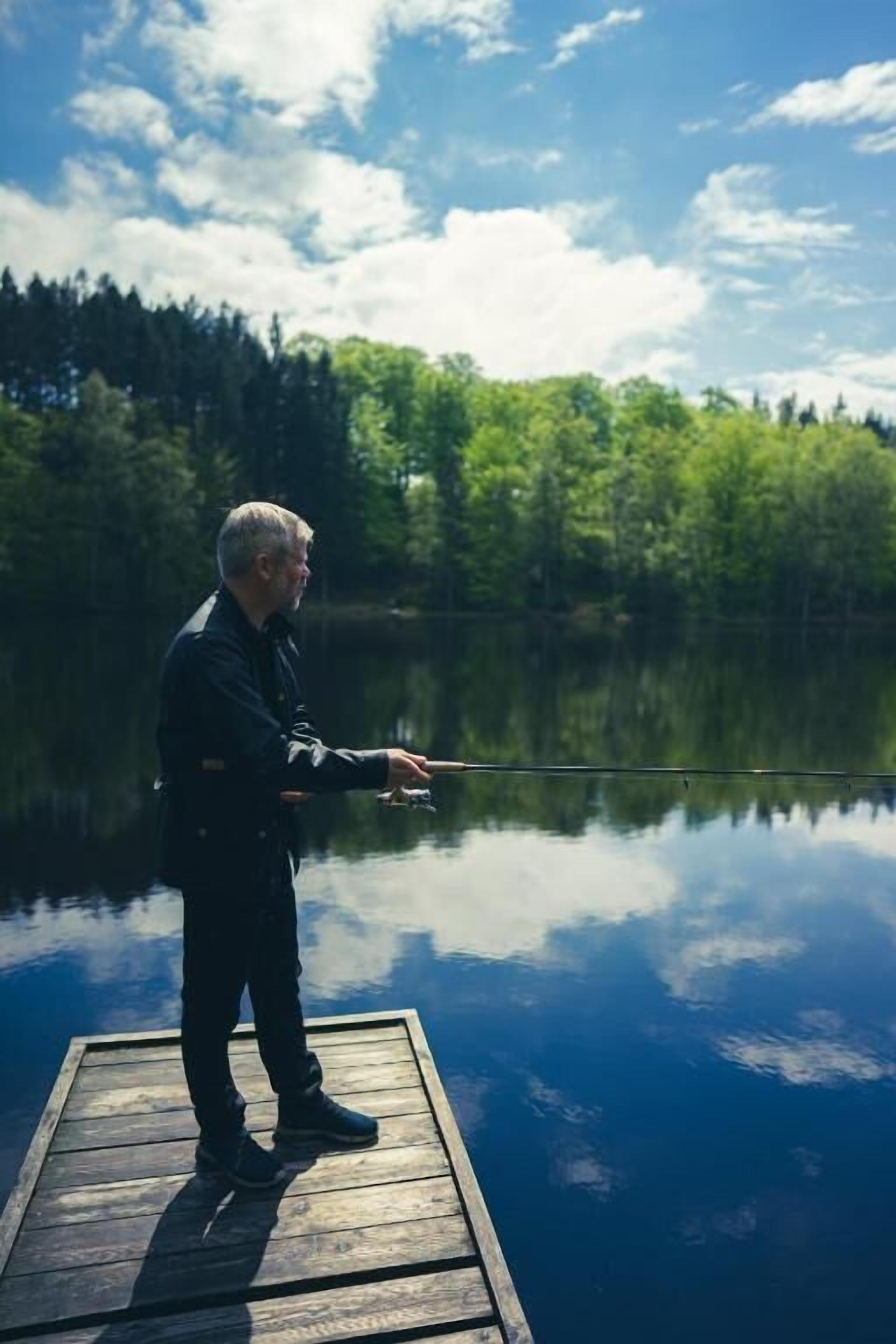 A person standing on a wooden dock extending into a calm lake, surrounded by dense green trees.