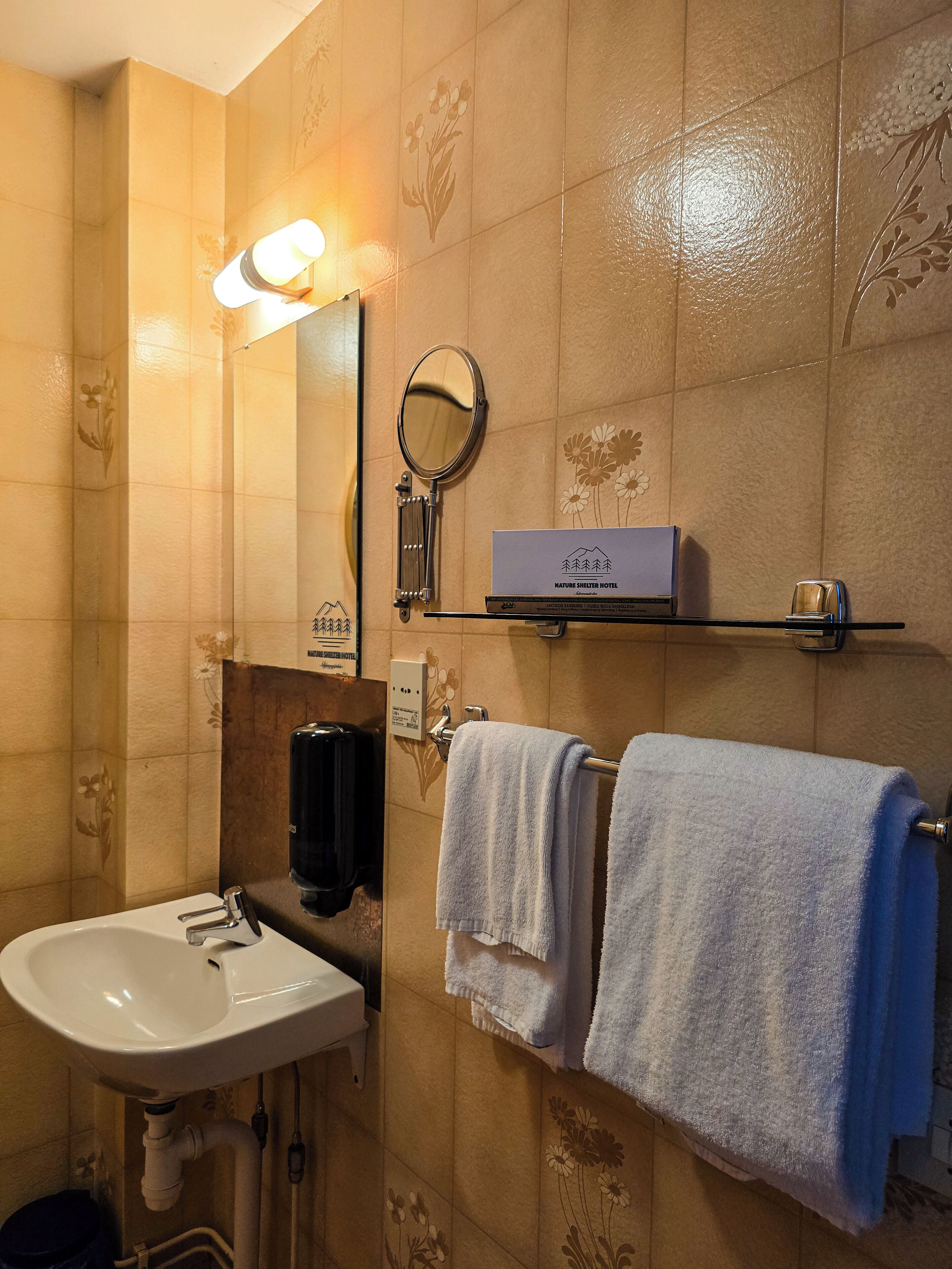 Ein Badezimmer mit Waschbecken, Spiegel, Handtuchhalter und einem Regal mit einer Box, beschriftet mit 'Nature Shelter Hotel'.