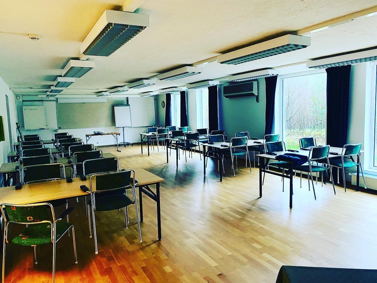 Ett rymligt konferensrum med flera rader av bord och stolar, stora fönster och en whiteboard.