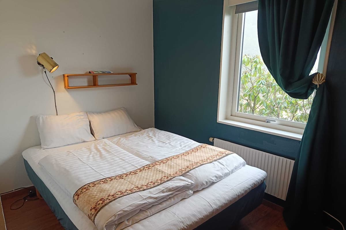 Cama bem arrumada com edredom branco e manta decorativa, mesa de cabeceira com abajur e janela com cortinas.