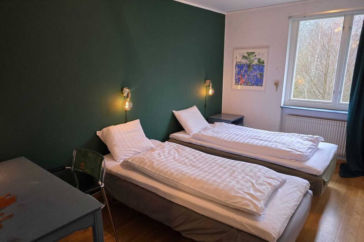 Quarto com duas camas de solteiro, uma mesinha, uma cadeira e janela com vista para árvores.