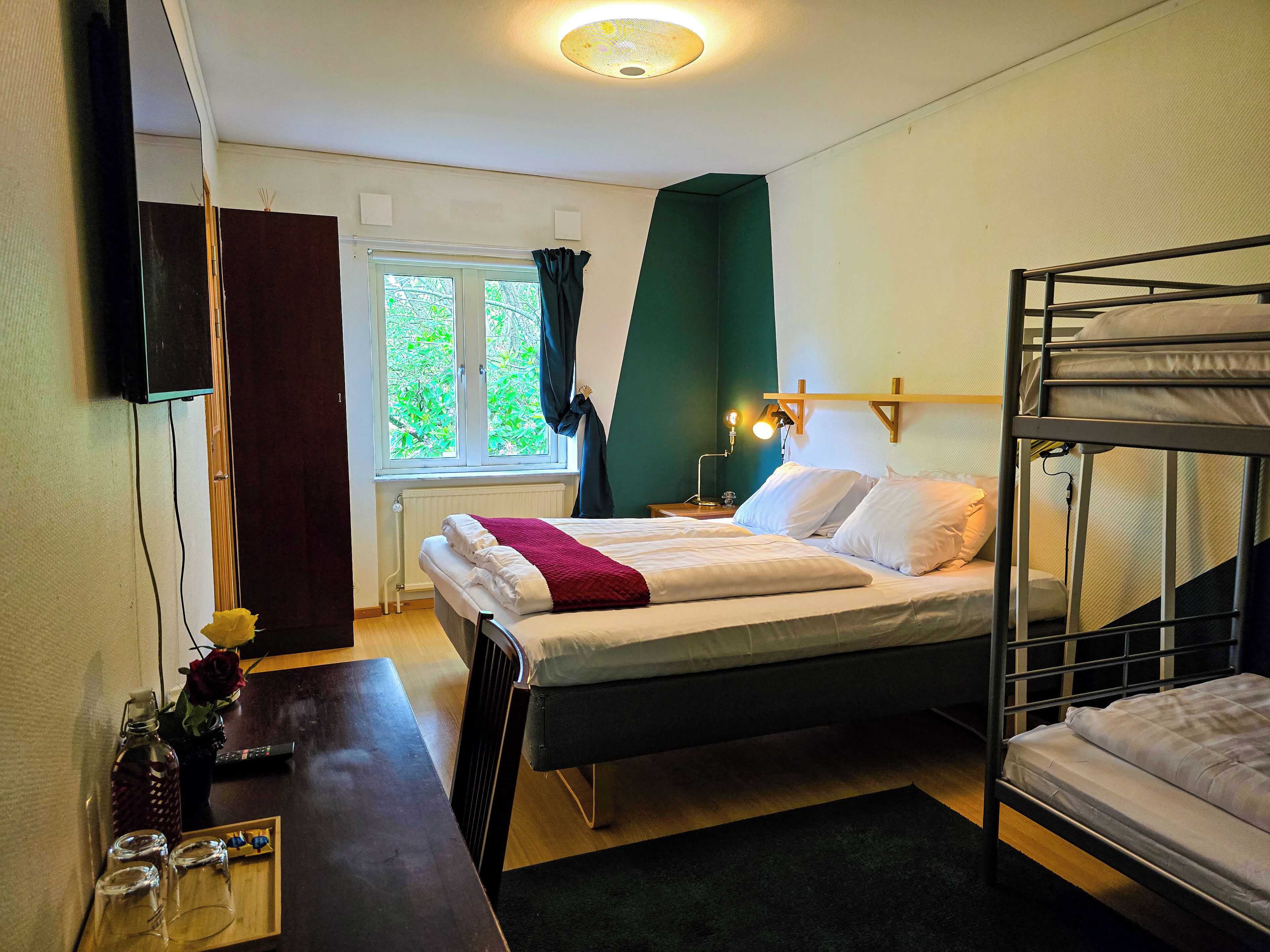 A bedroom with a bunk bed, a desk with a vase and glasses, a window with curtains, and a wall-mounted TV.