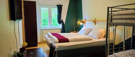 A bedroom with a bunk bed, a desk with a vase and glasses, a window with curtains, and a wall-mounted TV.