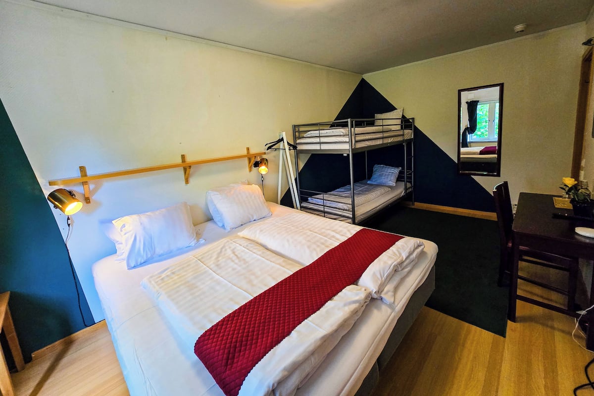 Quarto com beliche, escrivaninha e cadeira, uma cama com cobertor vermelho e um espelho na parede.