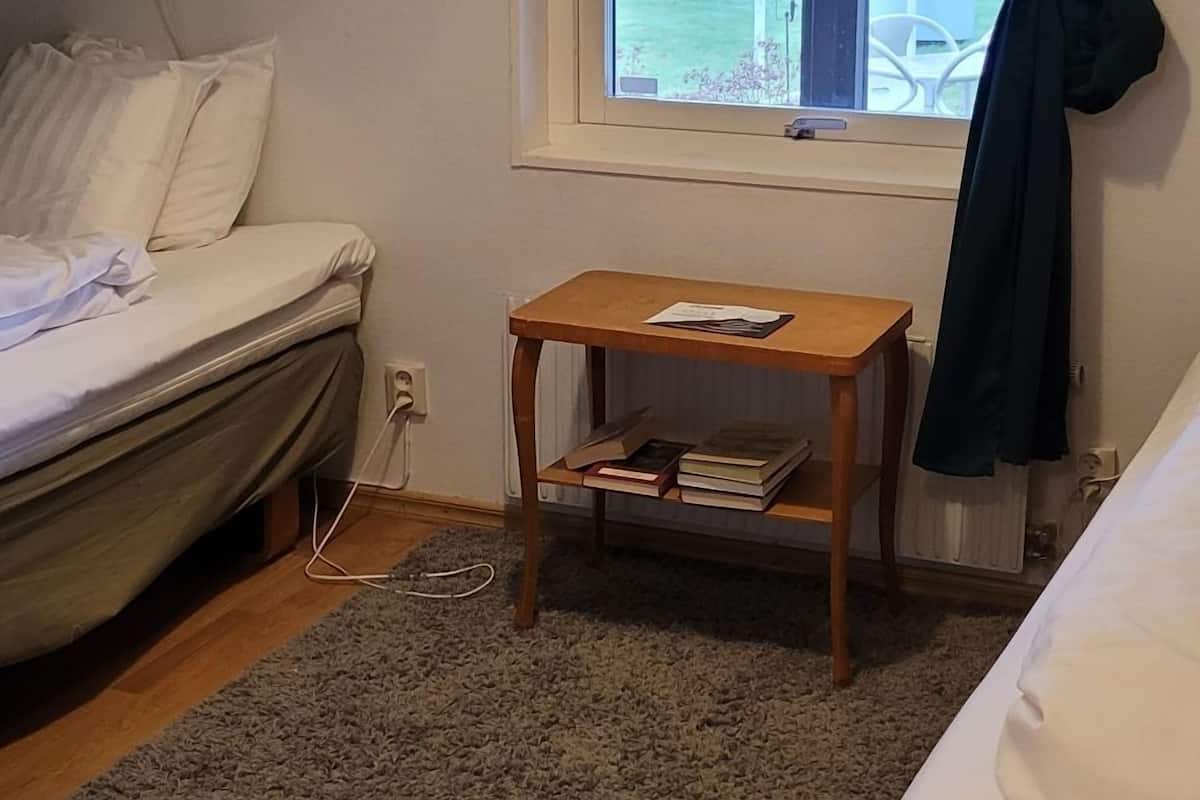 Quarto com duas camas, uma mesa de madeira pequena, uma janela com cortinas e uma luminária fixada na parede.