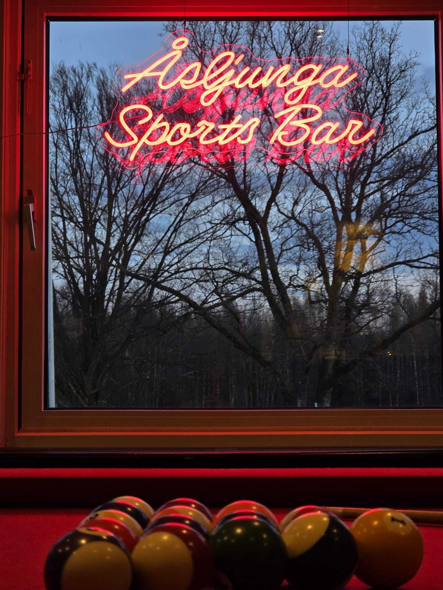 Une enseigne au néon indiquant « Asunga Sports Bar », avec, à travers une fenêtre, une vue sur des arbres sans feuilles.