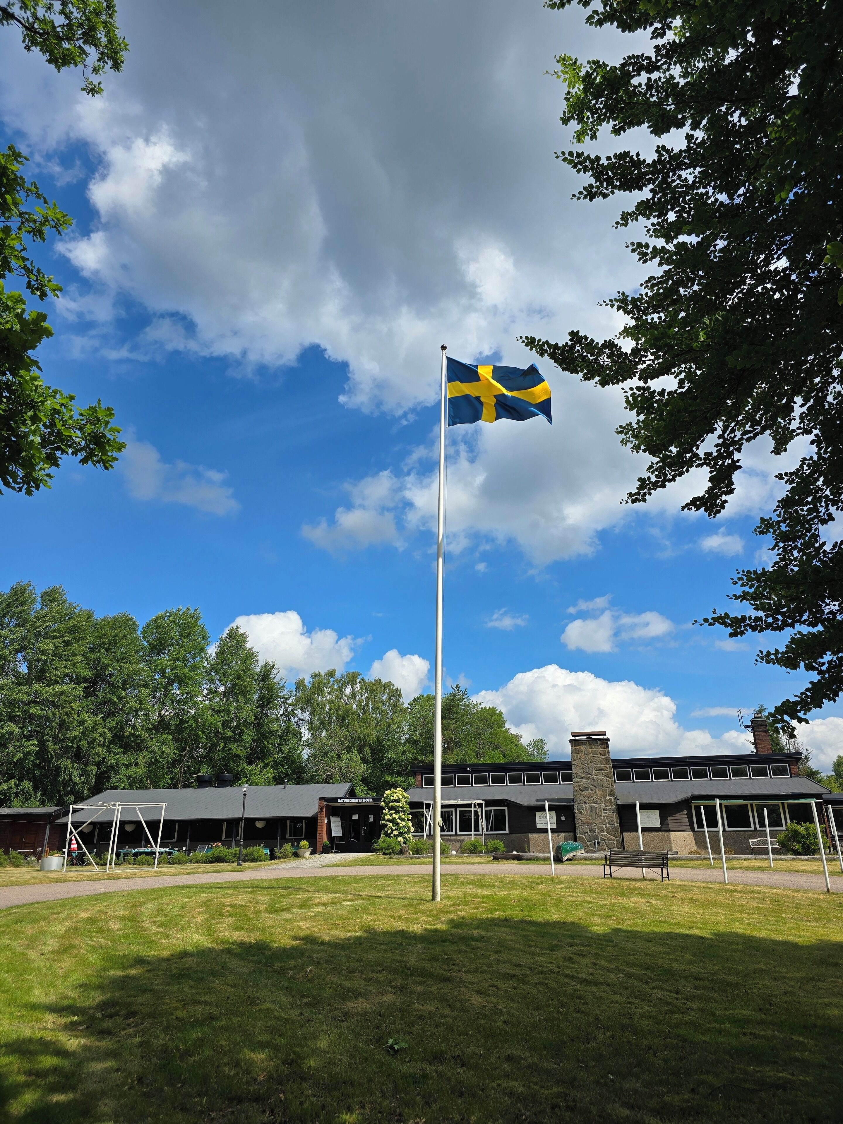 A flagpole with a Swedish flag, a building with a stone chimney, and a grassy area with a bench.