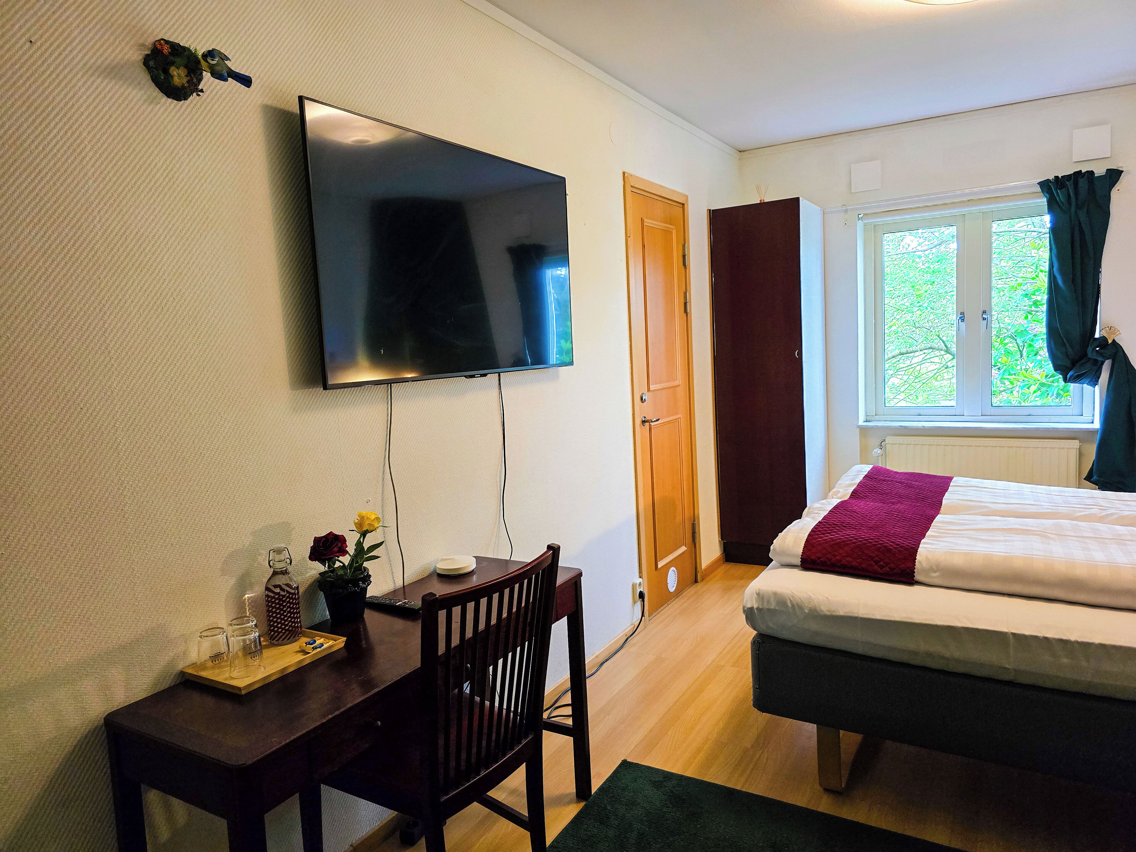 A bedroom with a bed, a desk with a chair, a TV mounted on the wall, and a window with curtains.
