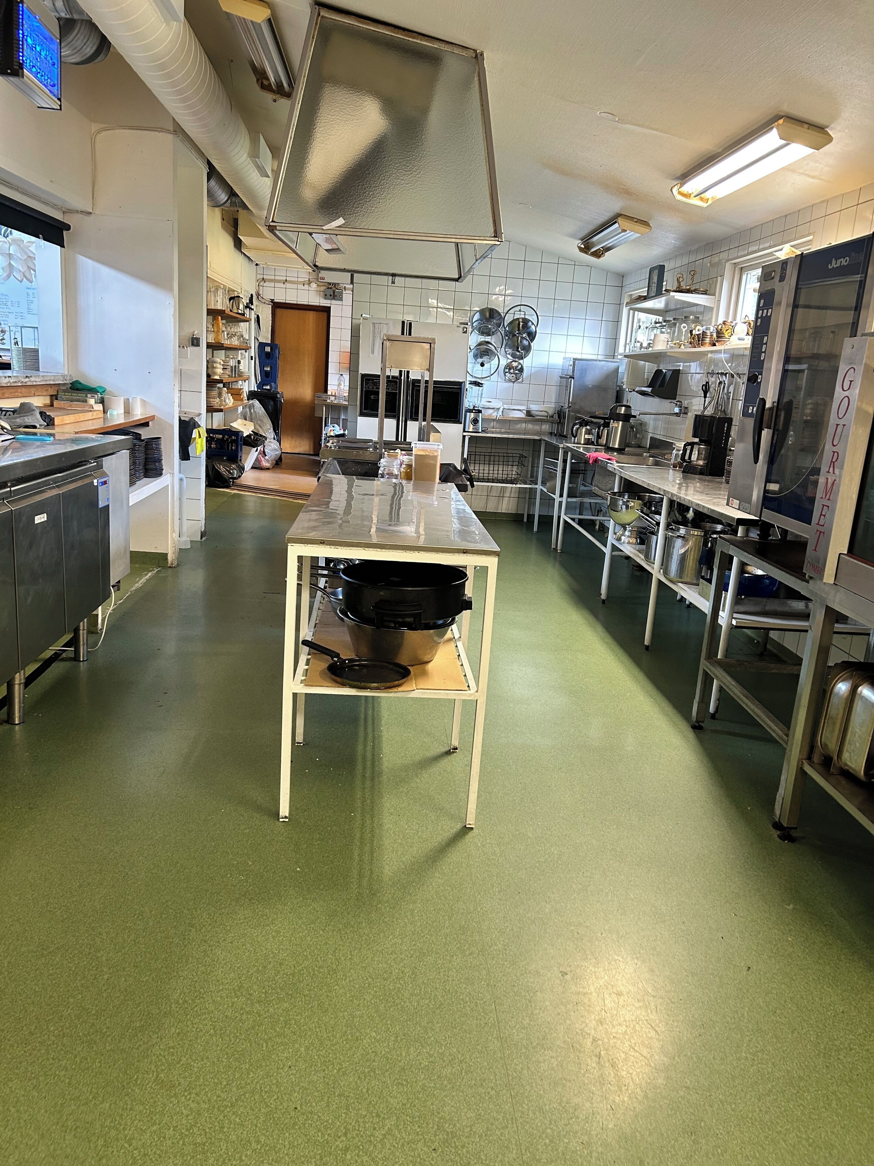 Ett rymligt professionellt kök med rostfria köksmaskiner, en central köksö och kakel på väggen bakom arbetsbänken.