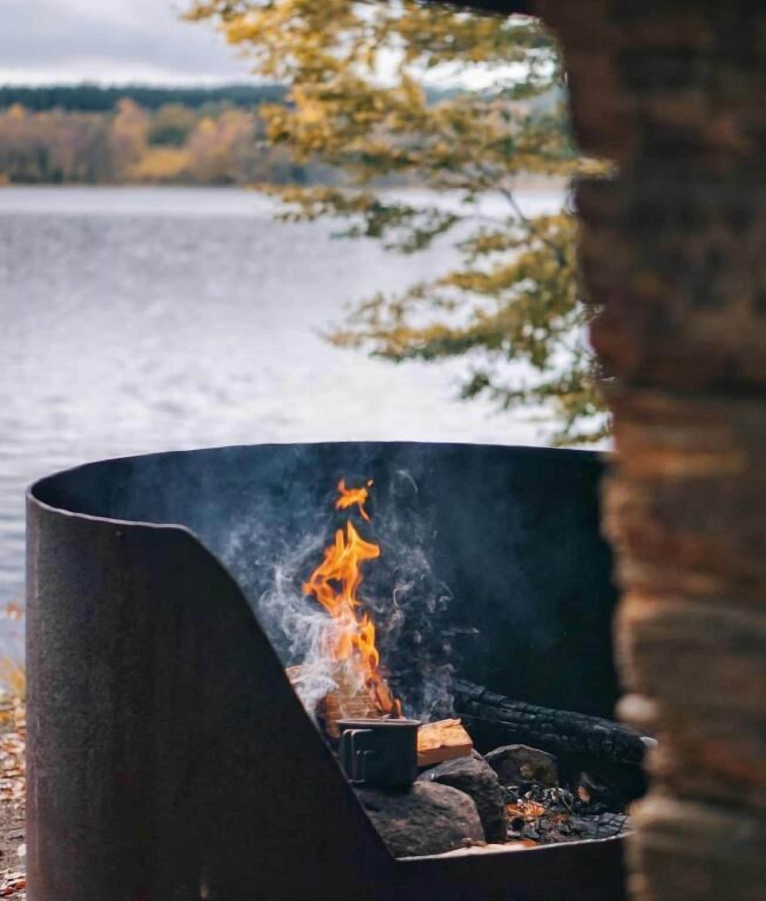 A metal fire pit with flames, surrounded by a natural setting with a lake and trees in the background.