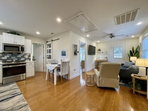 Uma cozinha moderna com armários brancos, micro-ondas e forno. Há uma área de jantar com mesa e cadeiras, e uma sala de estar com sofá e luminária.