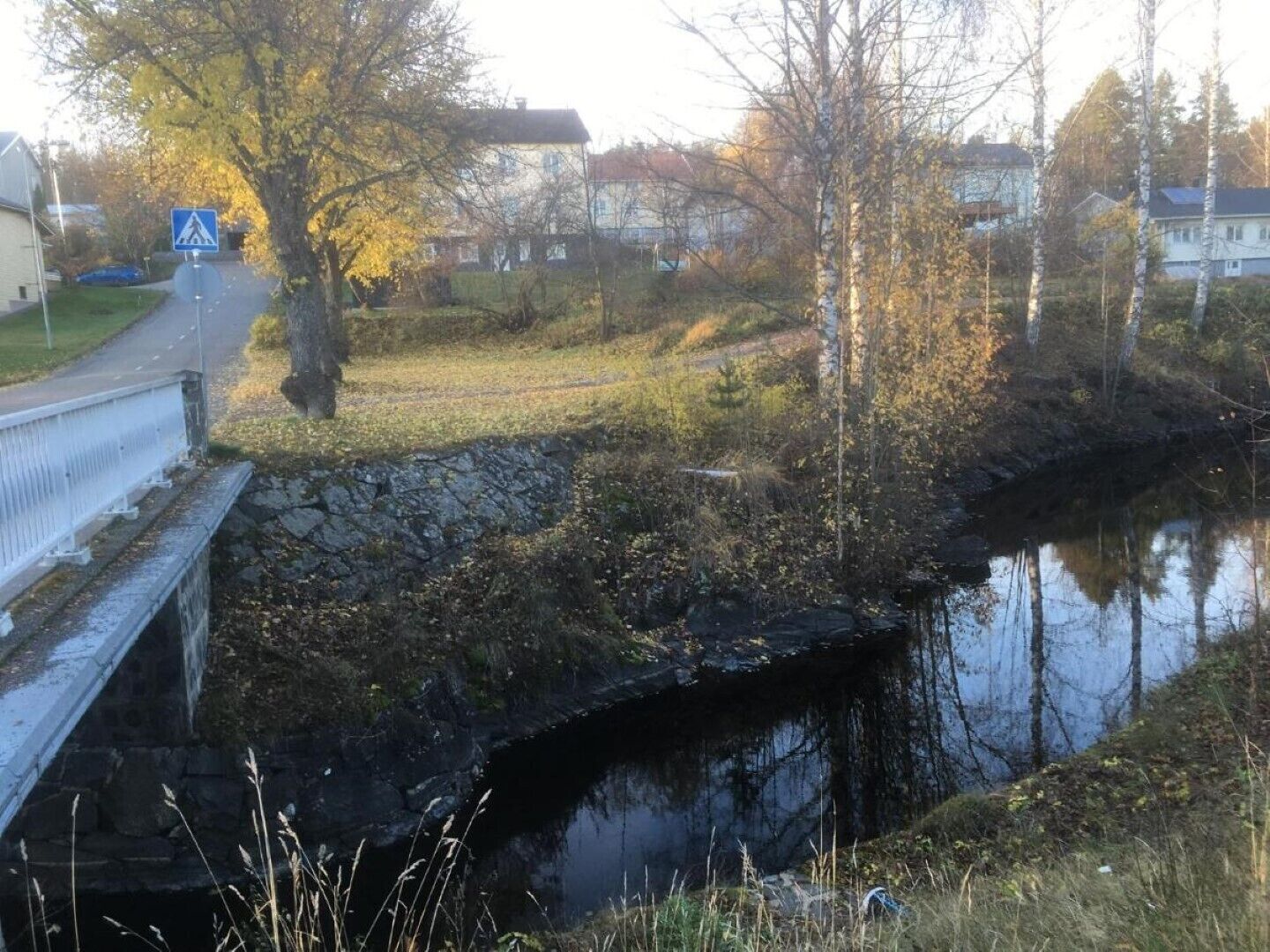 Kivisilta ylittää tyynen joen, ja sen yhteydessä on jalankulkijoiden ylityspaikka. Siltaa ympäröivät puut ja asuinrakennukset.