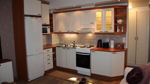 A compact kitchen with white cabinetry, a stove, a sink, and a microwave. There is a small dining area with a glass table and a purple cushion. - Cozy apartment in the city center close to nature and main attractions (Savonlinna)