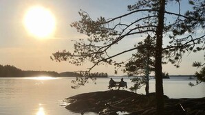 A serene lakeside scene with a clear view of the sun reflecting on the water, surrounded by trees and a rocky shoreline. - Cozy apartment in the city center close to nature and main attractions (Savonlinna)