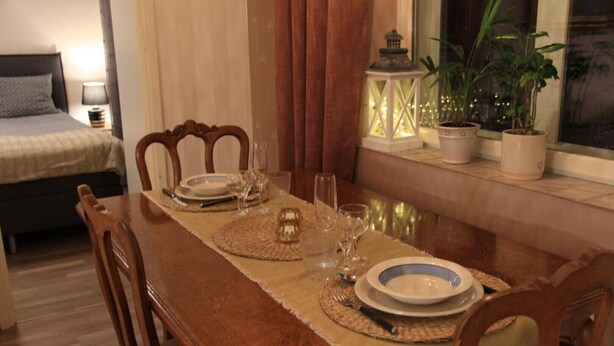 A dining area with a wooden table set for four, a bed with a headboard, and a window with plants and a lantern. - Cozy apartment in the city center close to nature and main attractions (Savonlinna)