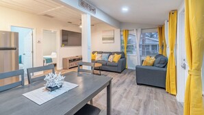 Ein modernes Wohnzimmer mit einem grauen Sofa, gelben Kissen, einem Esstisch mit Stühlen und einer Küchenzeile im Hintergrund.