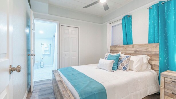 Ein Schlafzimmer mit einem hölzernen Kopfteil, einem Bett mit weißer Bettwäsche und Türkis-Akzenten, einem Nachttisch aus Holz und einem Deckenventilator.