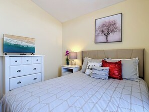 A bedroom with a bed, a dresser, a TV, a nightstand, a lamp, and a wall-mounted picture.