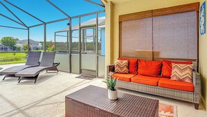 Een overdekt terras met een rietenbank en fauteuils, een bloempot en uitzicht op een huis en groen.