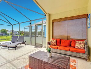 Een overdekt terras met een rietenbank en fauteuils, een bloempot en uitzicht op een huis en groen.
