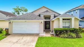 A residential house with a garage, a front door, and a small front yard with grass and shrubs. - Dream Weaver Villa® - Blooming Alamanda: Lakefront, Pool, Spa, Near Disney (Kissimmee)