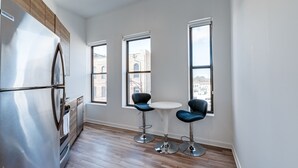 A modern kitchen with a stainless steel refrigerator, wooden cabinets, and a view of a building through large windows. There are two blue bar stools and a small round table. - Chicago Third Floor Studio by 747 Lofts (Chicago)