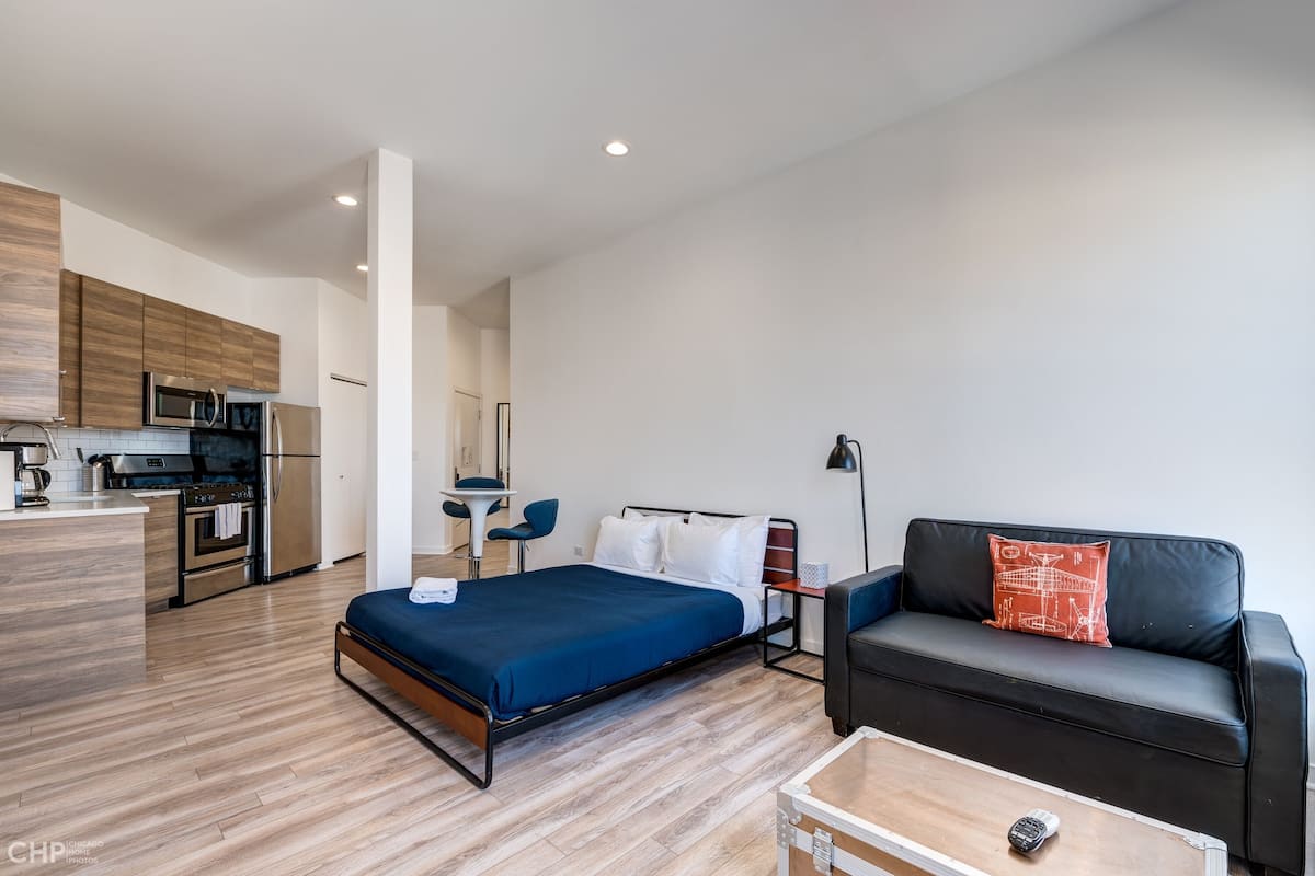 A modern hotel room with a bed, a sofa, a small table, a kitchenette, and a bathroom.