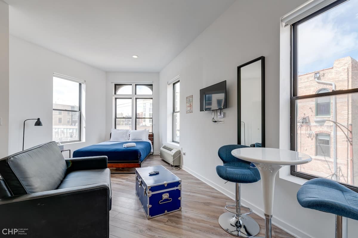A modern hotel room with a bed, a blue suitcase, a small round table, and a TV mounted on the wall.