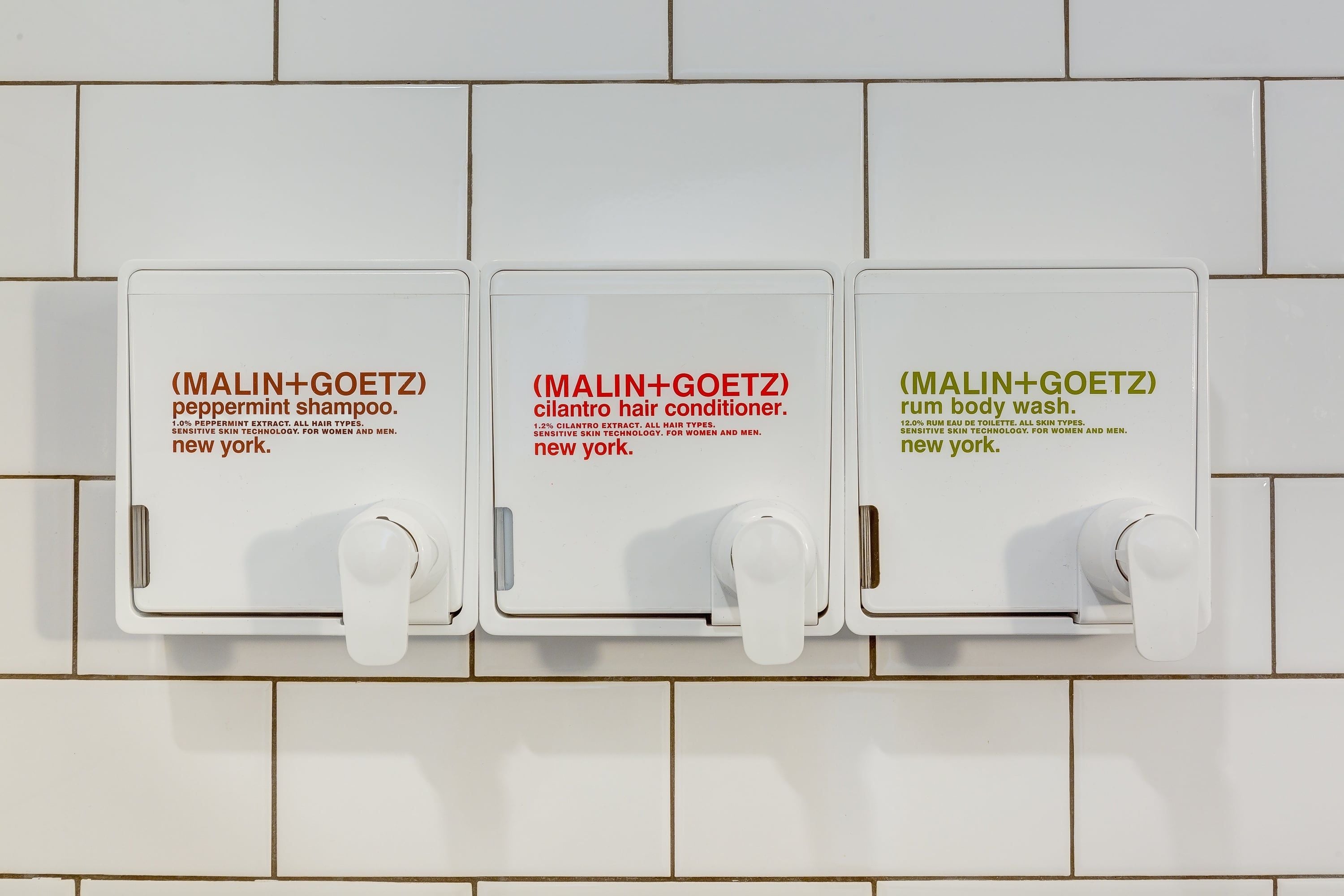 Three white dispensers on a tiled wall, each labeled with a product name and location.