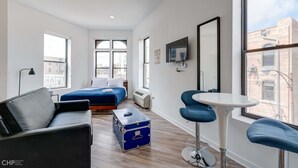 A modern hotel room with a bed, a sofa, a small table, a suitcase, and large windows offering city views. - Chicago Third Floor Studio by 747 Lofts (Chicago)
