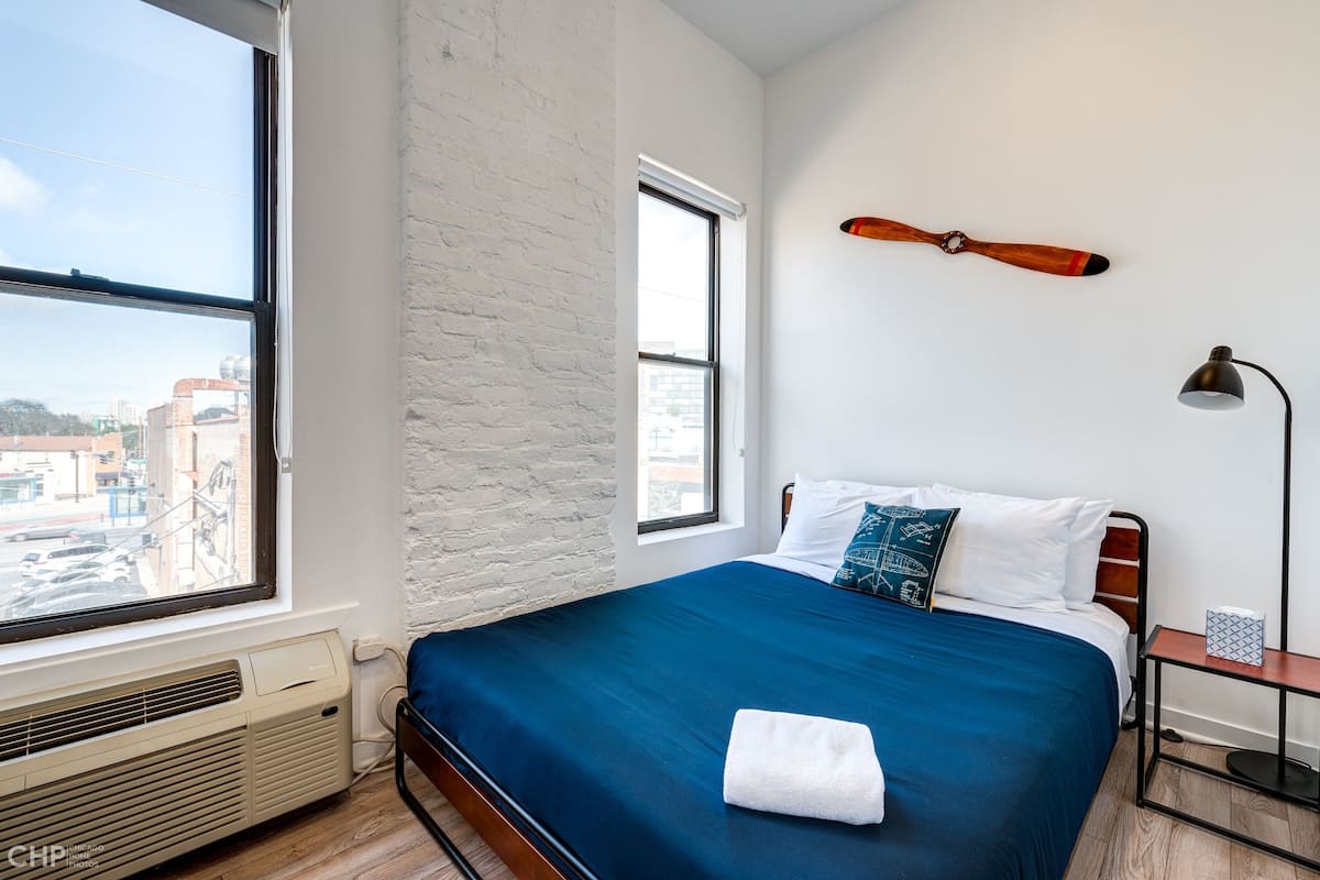 A bedroom with a large window, a bed with a blue bedspread, a white pillow, a wooden headboard, a wall-mounted lamp, and a small bedside table.