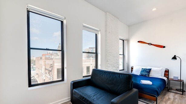 A modern bedroom with a bed, a black leather sofa, and large windows offering city views. - Chicago Third Floor Studio by 747 Lofts (Chicago)