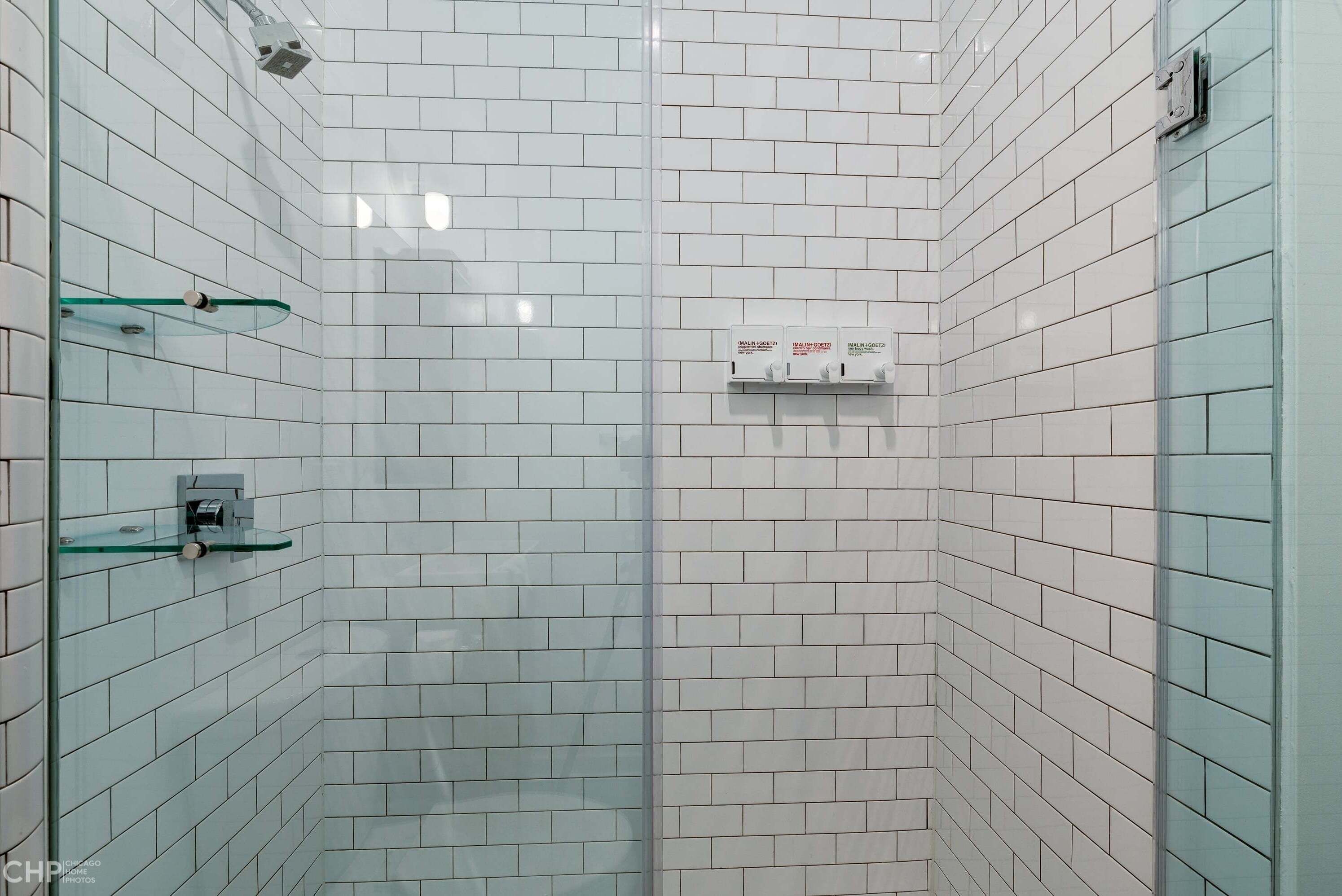 Ducha en esquina con azulejos blancos, estante de vidrio y panel de control.