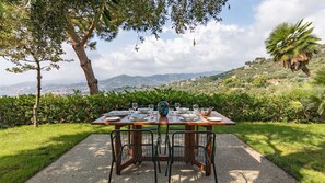Mesa de comedor al aire libre con sillas, platos y vasos, con vistas a un paisaje pintoresco de colinas y un pueblo lejano.