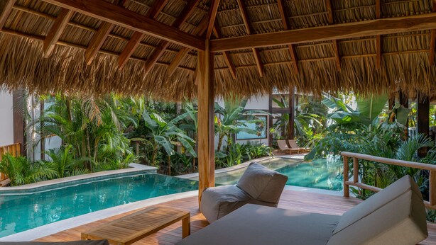 An indoor pool area with a thatched roof, wooden deck, and lounge chairs. - Blue Mist Hotel & Beach Club (Cóbano)