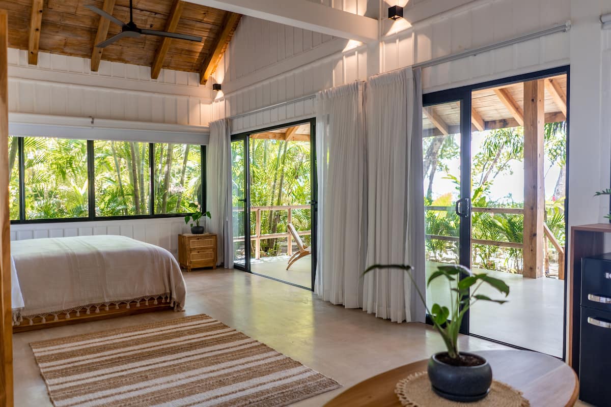 A spacious bedroom with a large bed, a wooden nightstand, a potted plant, and a view of lush greenery through large sliding glass doors.