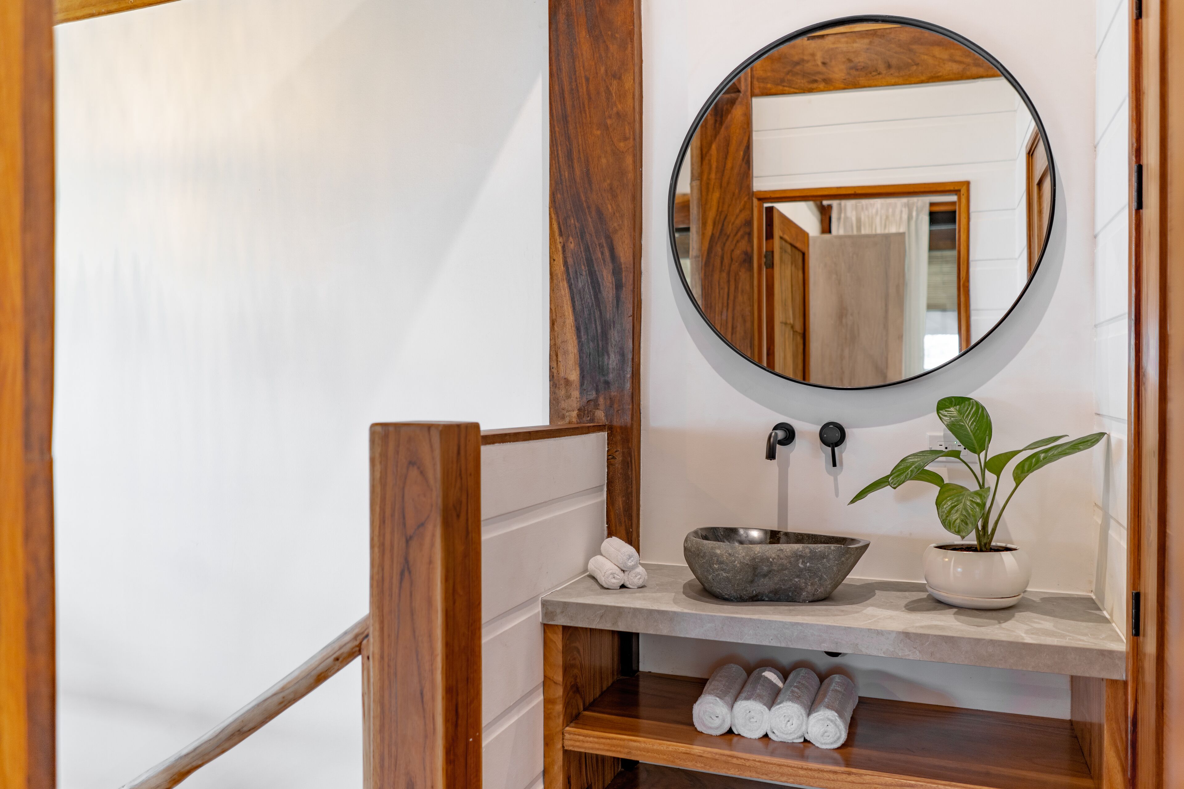 A bathroom with a wooden vanity, a round mirror, a stone basin, a potted plant, and neatly rolled towels on a shelf.