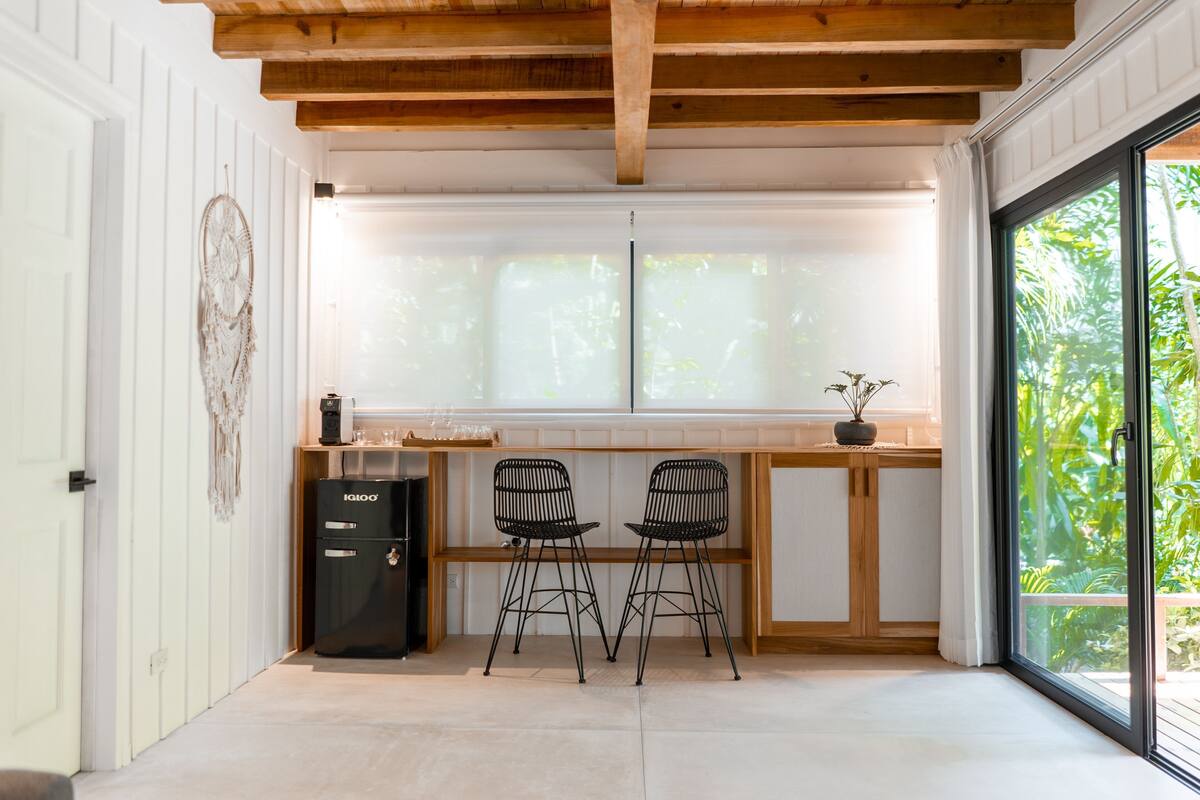A kitchen with a refrigerator, two chairs, a wooden table, and a large window with a view of greenery.