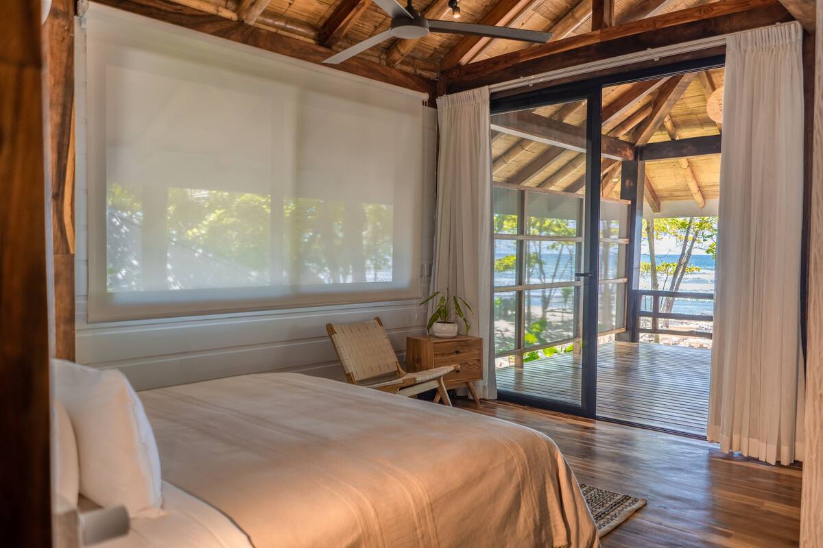 A bedroom with a large bed, a wooden ceiling, a sliding glass door to a balcony, and a view of the beach.