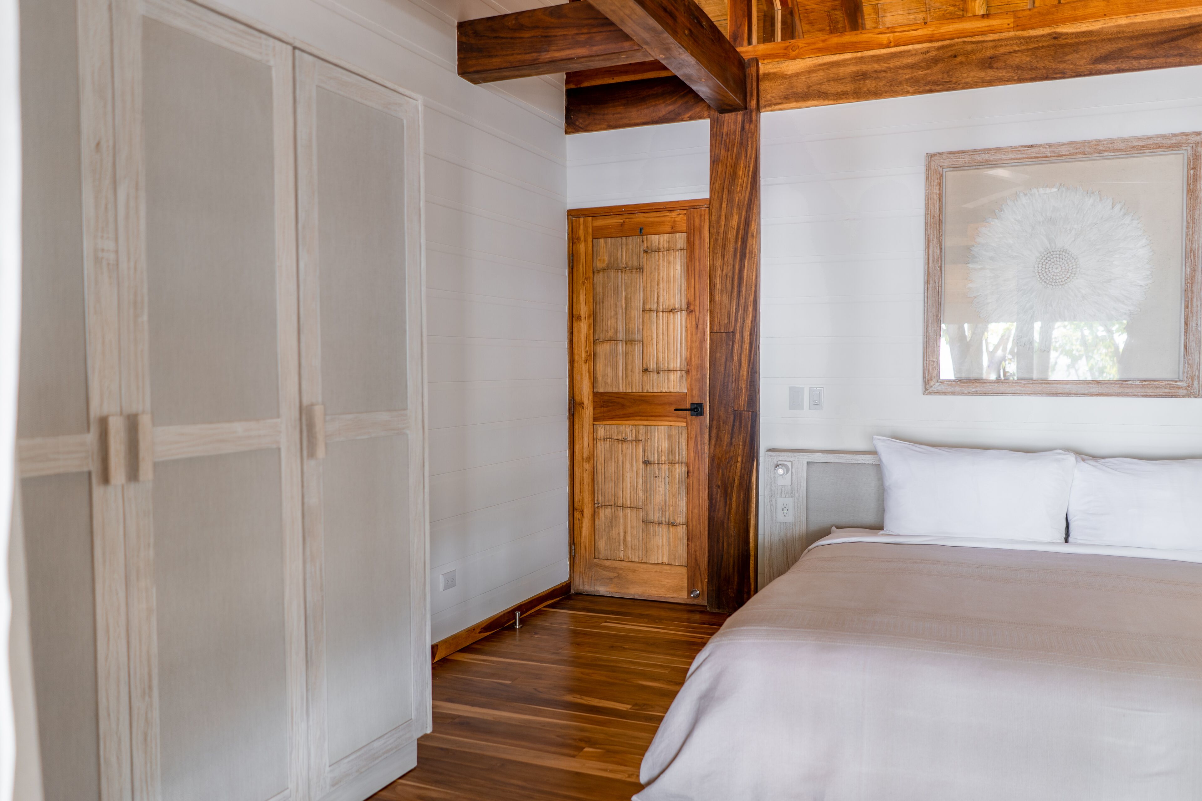 A bedroom with a wooden floor, a bed with white linens, a wooden door, and a framed picture on the wall.