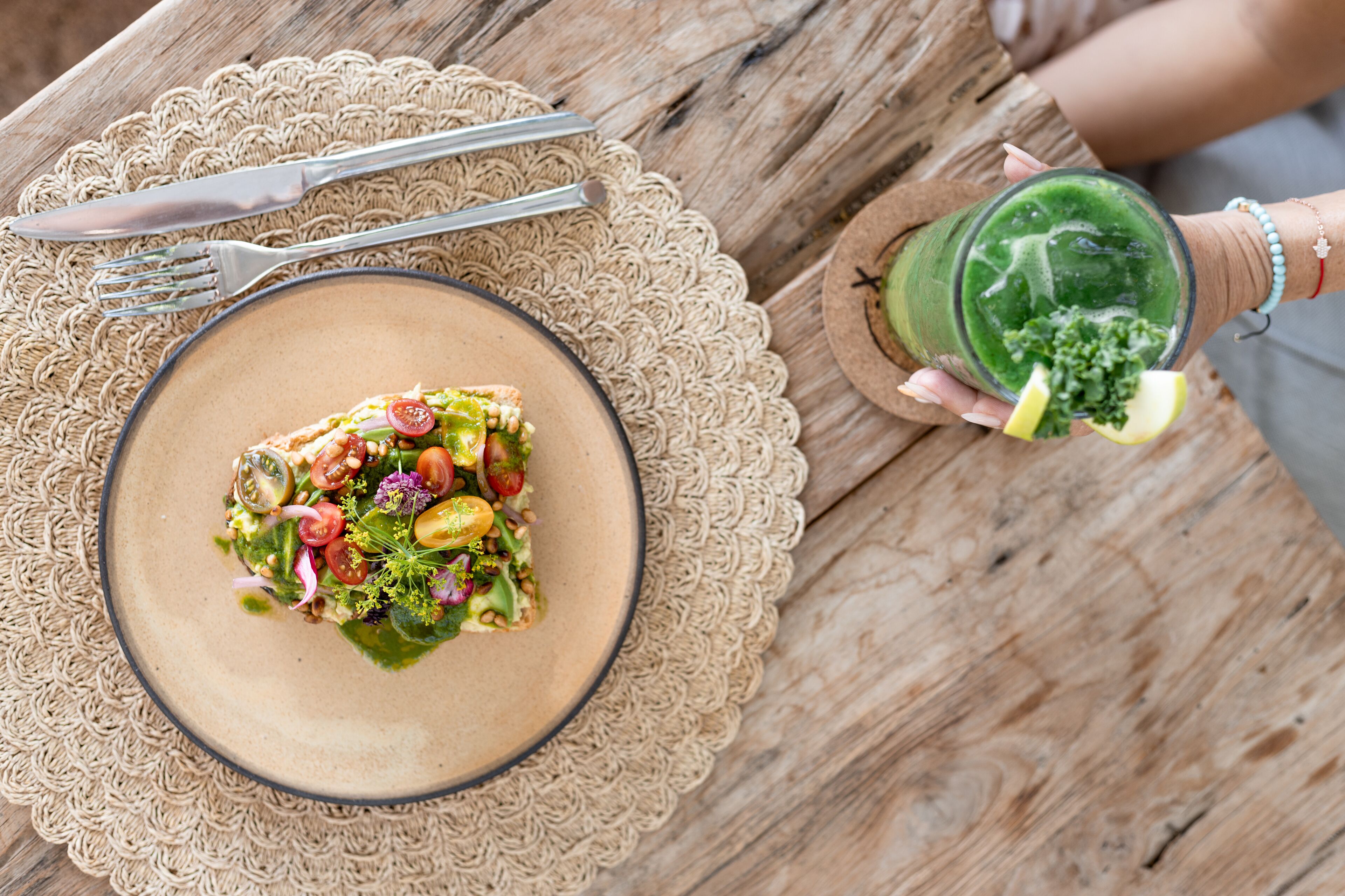 A plate of salad on a woven placemat, a fork and knife on a wooden table, and a glass of green drink with a lemon slice.