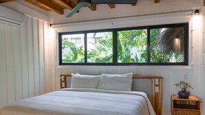 A bedroom with a large bed, a window with a view of greenery, and a small wooden nightstand with a plant. - Blue Mist Hotel & Beach Club (Cóbano)