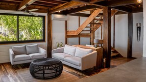 A spacious living area with a high ceiling, wooden beams, a large window with a view of greenery, two sofas, a round black wicker table, and a staircase. - Blue Mist Hotel & Beach Club (Cóbano)