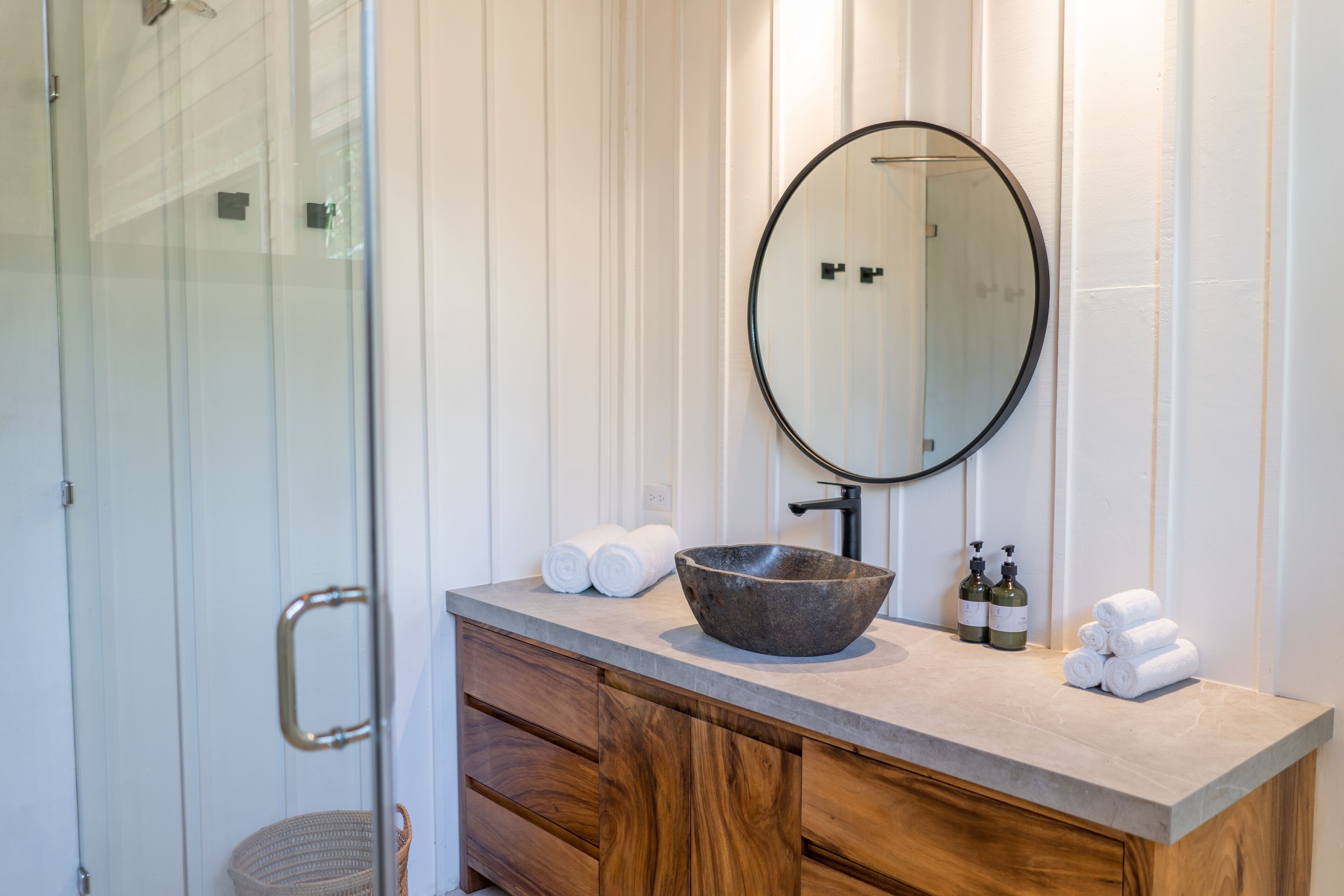 A bathroom with a wooden vanity, a large round mirror, and a glass shower enclosure.